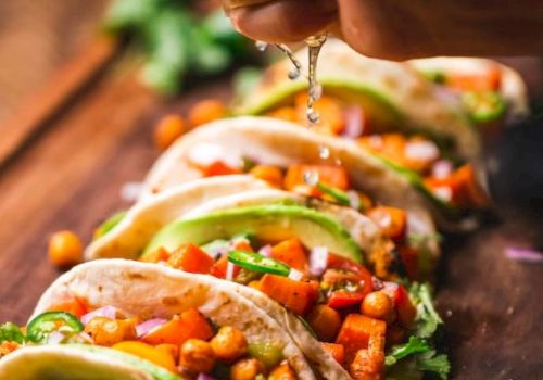 The image shows a hand squeezing lime juice onto a row of colorful tacos filled with vegetables, chickpeas, and garnishes on a wooden board.