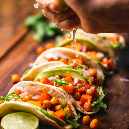 The image shows a hand squeezing lime juice onto a row of colorful tacos filled with vegetables, chickpeas, and garnishes on a wooden board.