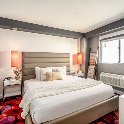 A modern hotel bedroom with a padded headboard, white bedding, two red lamps, a colorful red patterned carpet, and a large window with a view.