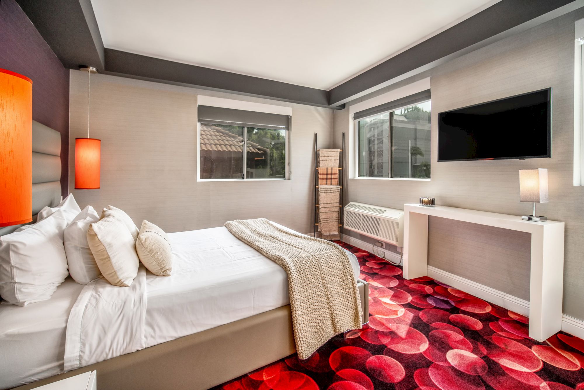 A modern bedroom with a bed, two windows, a TV, and stylish decor, featuring a bold patterned carpet and ambient lighting.