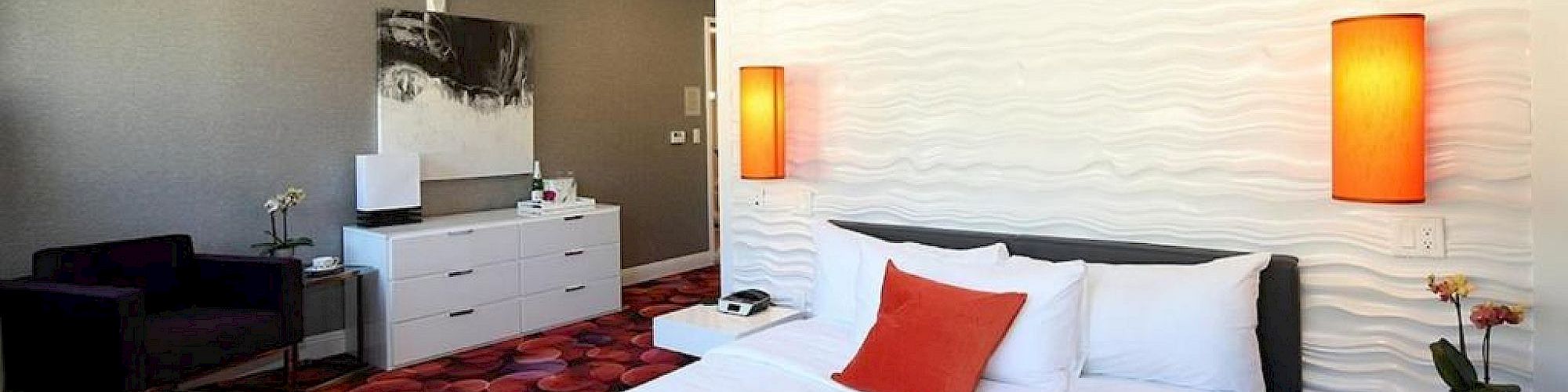 A modern hotel room features a double bed with red and white bedding, vibrant carpet, white furniture, and ambient lighting with orange lamps.