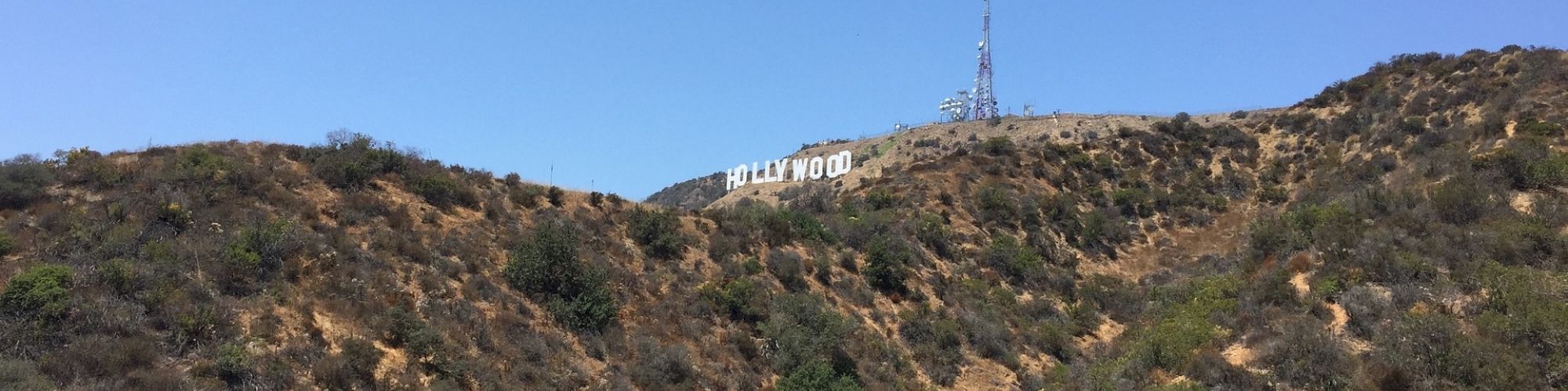 The image shows a distant view of the Hollywood sign situated on a hillside covered with sparse vegetation under a clear blue sky.