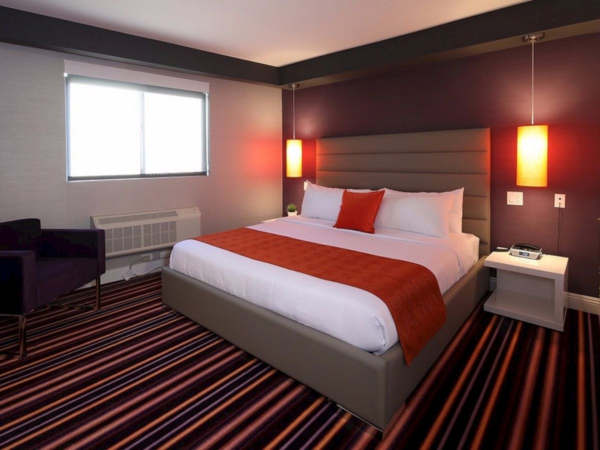 A modern hotel room features a king-sized bed with orange accents, striped carpet, an armchair, two pendant lights, and a window.