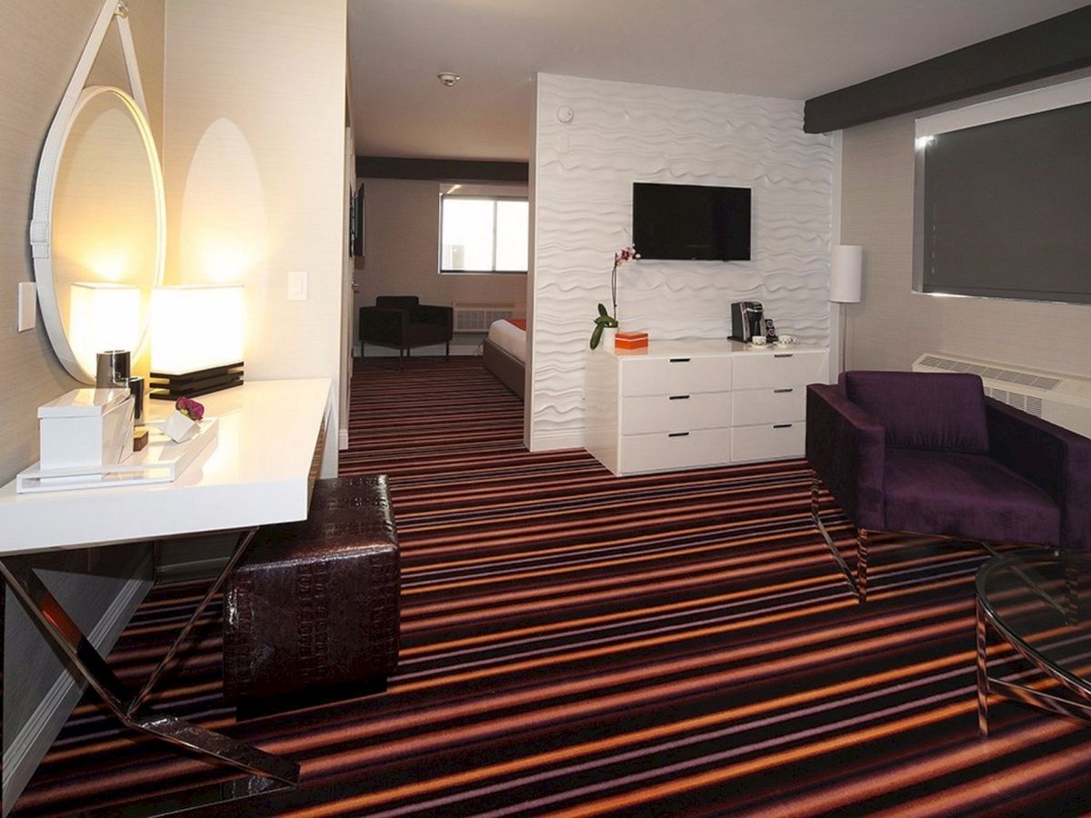 A modern hotel room with striped carpet, a vanity, purple chairs, TV, dresser, and a visible cozy bed area in the back.