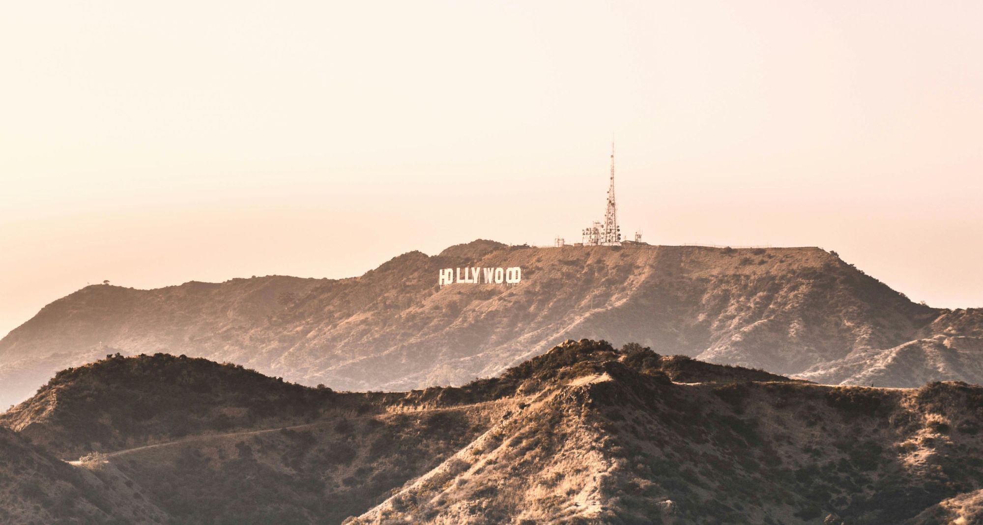 This image shows the iconic Hollywood sign on a hill, with a television tower nearby, set against a backdrop of rugged terrain and a clear sky.