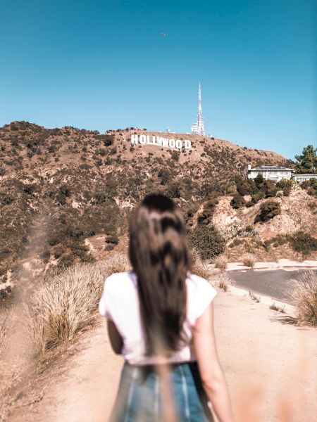 A person stands on a dirt path looking towards the iconic Hollywood sign on a hill under a clear blue sky.