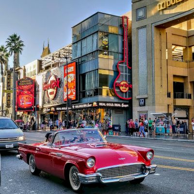 The image features a red classic convertible driving on a city street lined with buildings, billboards, and a guitar-shaped sign, in what seems to be a busy area.