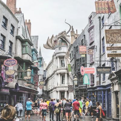 A busy street scene in a themed area with storefronts, signs, and a large dragon on a building, filled with visitors and tourists strolling around.