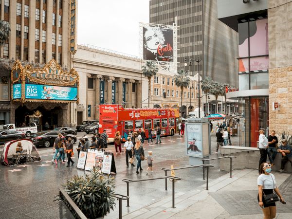 A bustling city street with pedestrians, tour buses, and classic theater marquees, set against a backdrop of tall buildings and busy activity.