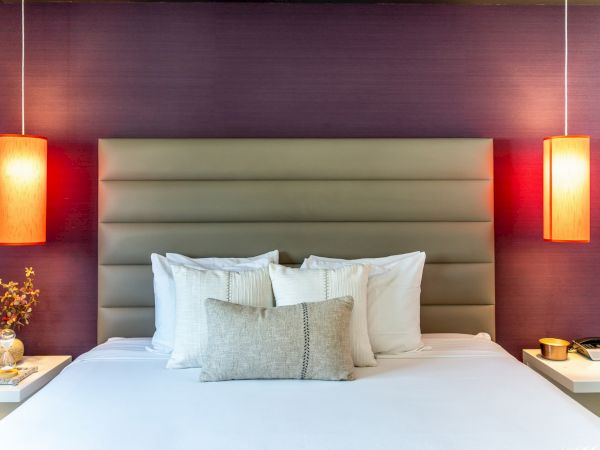 A modern bedroom with a bed, pillows, and two hanging orange lamps on either side, set against a purple wall.