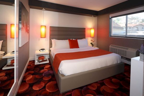 A modern bedroom with a large bed, red and white bedding, bedside tables, and orange pendant lights. The room has a window and colorful carpet.