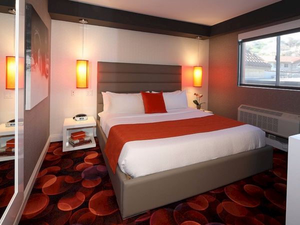 A modern bedroom with a large bed, red and white bedding, bedside tables, and orange pendant lights. The room has a window and colorful carpet.