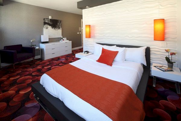 A modern bedroom has a bed with orange and white bedding, red lamps, a white dresser, a chair, and artwork on the wall.