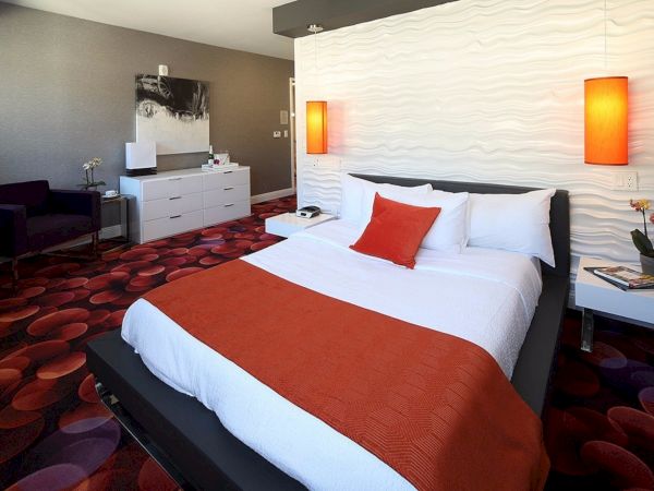 A modern bedroom has a bed with orange and white bedding, red lamps, a white dresser, a chair, and artwork on the wall.