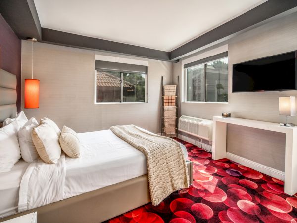 A modern bedroom with a bed, vibrant carpet, wall-mounted TV, and large windows, featuring neutral tones and bright accents.