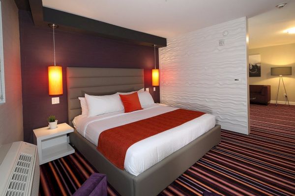 A modern hotel room with a large bed, two red pendant lights, a striped carpet, and a white textured wall.