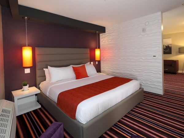 A modern hotel room with a large bed, two red pendant lights, a striped carpet, and a white textured wall.