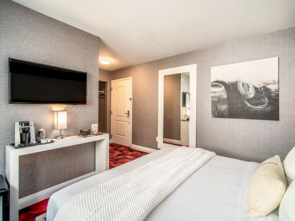 A modern hotel room with a bed, wall-mounted TV, a desk with a lamp, coffee maker, and wall art. Neutral colors create a cozy atmosphere.