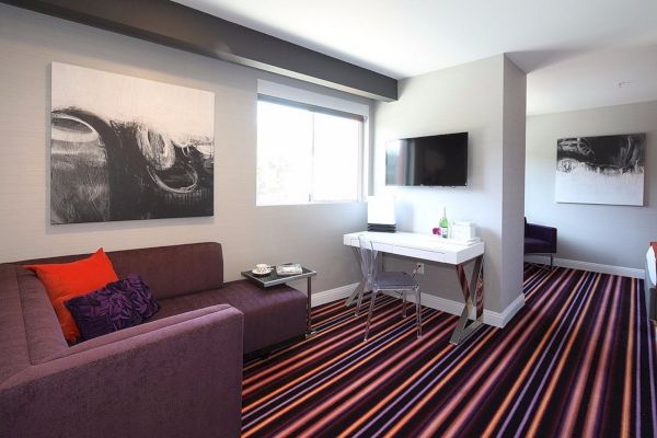 A modern lounge with a purple sofa, vibrant striped carpet, abstract wall art, a TV, a white desk with a chair under a window, and tea set on a side table.