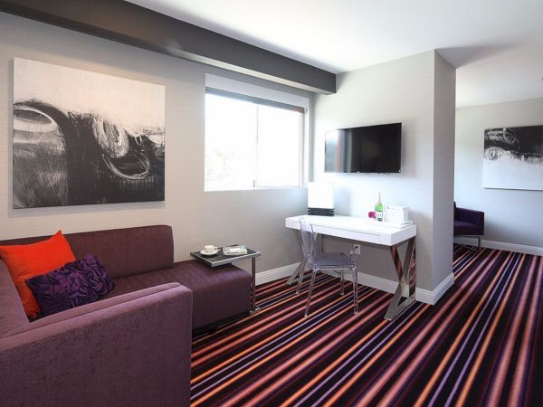 A modern lounge with a purple sofa, vibrant striped carpet, abstract wall art, a TV, a white desk with a chair under a window, and tea set on a side table.