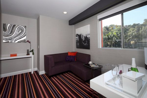 A modern room with striped flooring, artwork, a purple couch, a window with a view, a small table, and a desk holding a wine bottle and glasses.