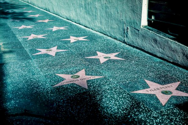 The image shows a sidewalk with pink stars on the ground, resembling the Hollywood Walk of Fame. The stars have names and are set against a grey background.