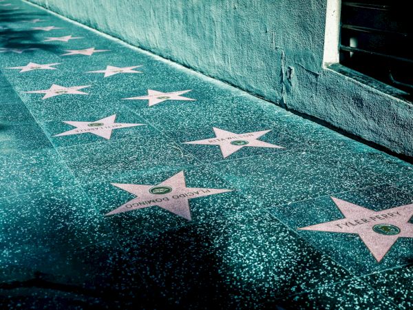 The image shows a sidewalk with pink stars on the ground, resembling the Hollywood Walk of Fame. The stars have names and are set against a grey background.