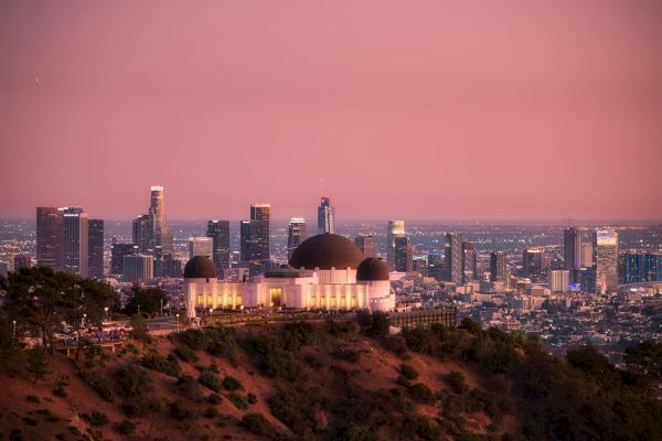 An observatory sits atop a hill, with a sprawling cityscape and skyscrapers in the background under a pink sky during sunset, ending the sentence.