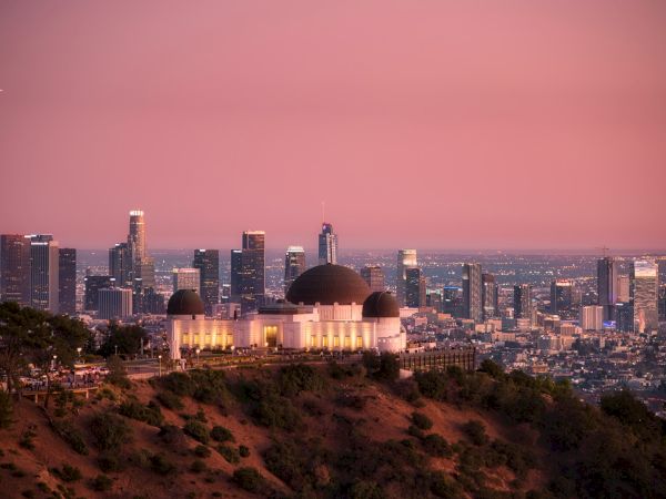 An observatory sits atop a hill, with a sprawling cityscape and skyscrapers in the background under a pink sky during sunset, ending the sentence.