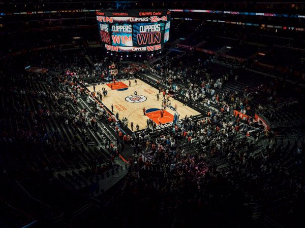 An indoor basketball arena showing a "CLIPPERS WIN" on the scoreboard with players and fans on the court and in the stands.