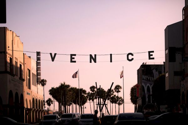 The image depicts a street in Venice, California, with a sign hanging between buildings that spells 