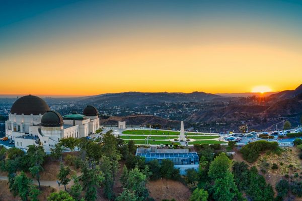 The image shows an iconic observatory building at sunset, set on a hill with a panoramic view of a sprawling cityscape and lush greenery surrounding it.