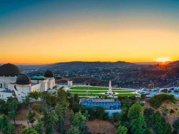 The image shows an iconic observatory building at sunset, set on a hill with a panoramic view of a sprawling cityscape and lush greenery surrounding it.