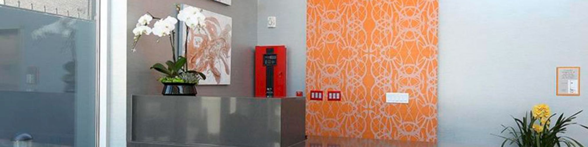 A modern reception area with artistic wall decor, a textured front desk, hanging lights, and plants. An emergency alarm is mounted on the wall.