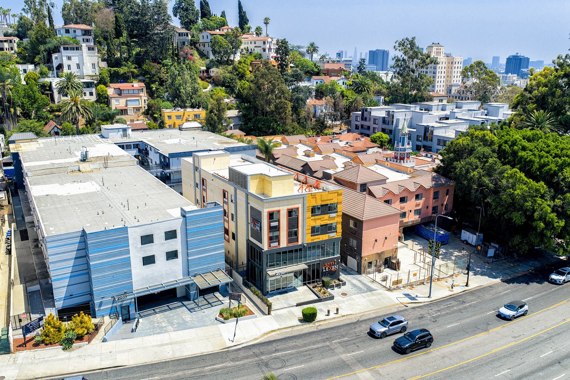 Aerial view of an urban area with a mix of modern buildings, houses, and greenery along a main road.