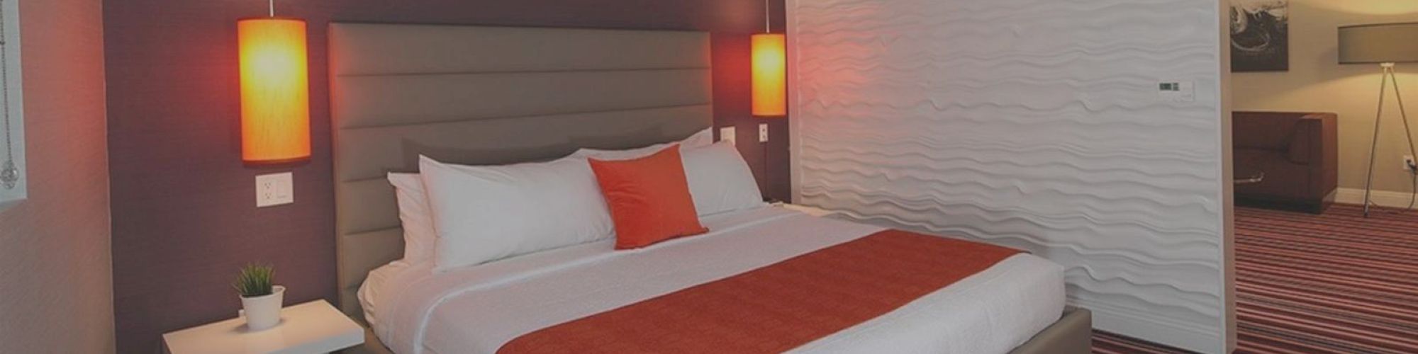 The image shows a modern bedroom with a double bed, orange accents, hanging lights, and a nightstand with a potted plant.