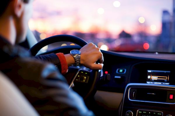 A person is driving a car, with their hand on the steering wheel, looking at the road ahead as dusk sets in outside the vehicle.