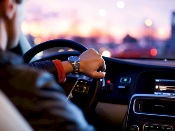 A person is driving a car, with their hand on the steering wheel, looking at the road ahead as dusk sets in outside the vehicle.