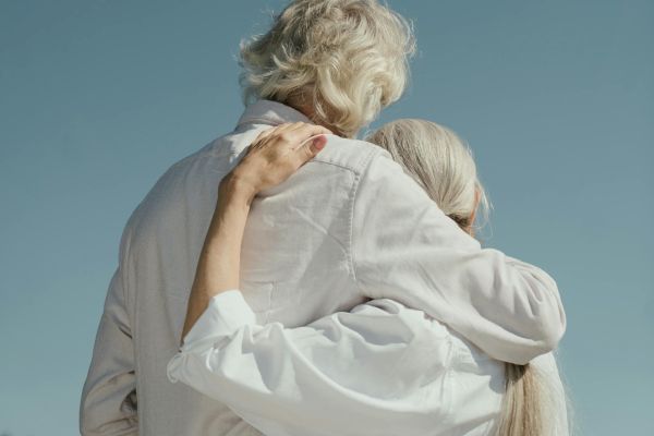 Two elderly individuals with white hair embrace tenderly in the foreground, set against a clear blue sky in the background.