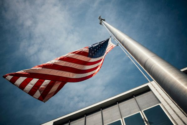 The image shows an American flag waving on a flagpole against a blue sky, with part of a building visible in the background.