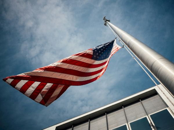 The image shows an American flag waving on a flagpole against a blue sky, with part of a building visible in the background.