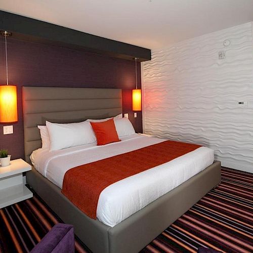 A modern hotel room features a comfortable bed with orange accents, striped carpet, purple walls, and contemporary lighting.