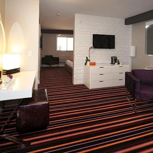 This image shows a modern hotel room with a striped carpet, a vanity desk, a chair, a bed in the background, and a TV mounted on the wall.