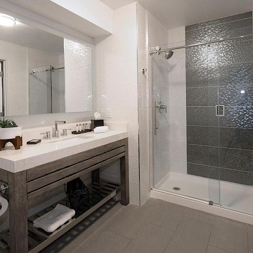 The image shows a modern bathroom with a large mirror, a spacious vanity with towels, and a glass-enclosed shower with tiled walls.