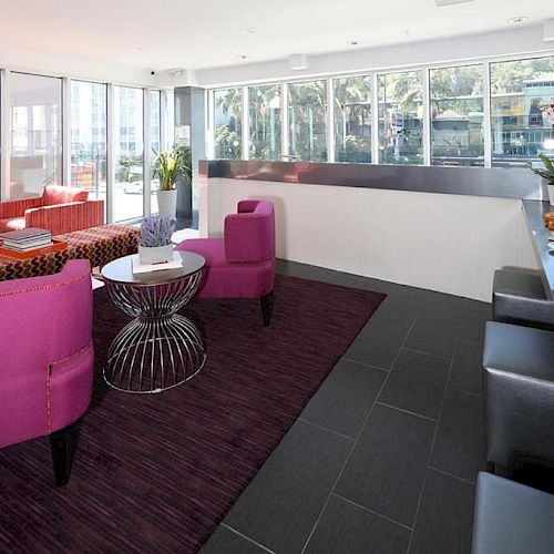 A modern lounge area with pink chairs, glass-topped tables, a counter with snacks, and large windows letting in natural light, ending the sentence.