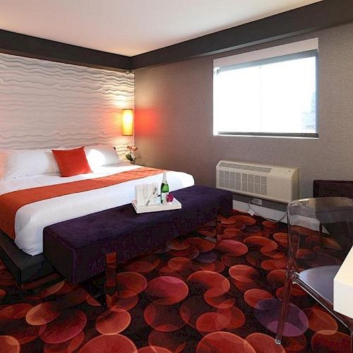 A modern hotel room features a king-size bed with red accents, a unique carpet pattern, ambient lighting, and a work desk by the window, all ending the sentence.