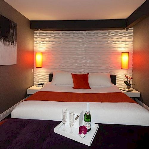 A modern hotel room with a double bed, red and white bedding, pendant lights, wall art, and a tray with champagne and glasses on the foot of the bed.