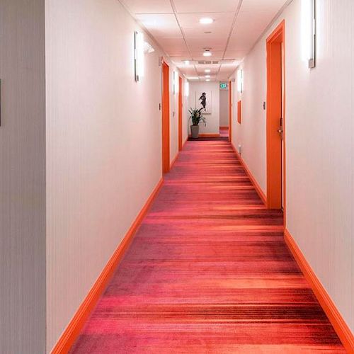 A hotel hallway with a pink and red carpet, light-colored walls, and illuminated signs for rooms on the left side of the corridor, ending the sentence.