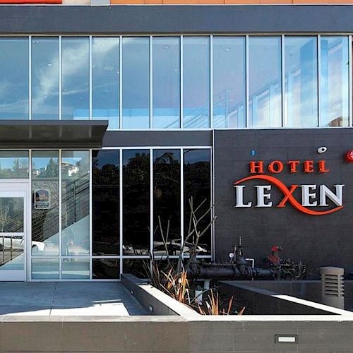 The image shows the front entrance of Hotel Lexen, with the number 2018 visible, featuring modern glass architecture and the hotel sign on the right.