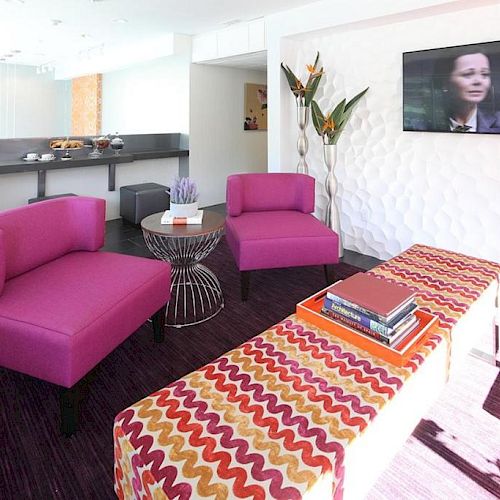 A modern lounge with purple chairs, a patterned bench, coffee table, TV, plants, and a counter with dishes in the background.